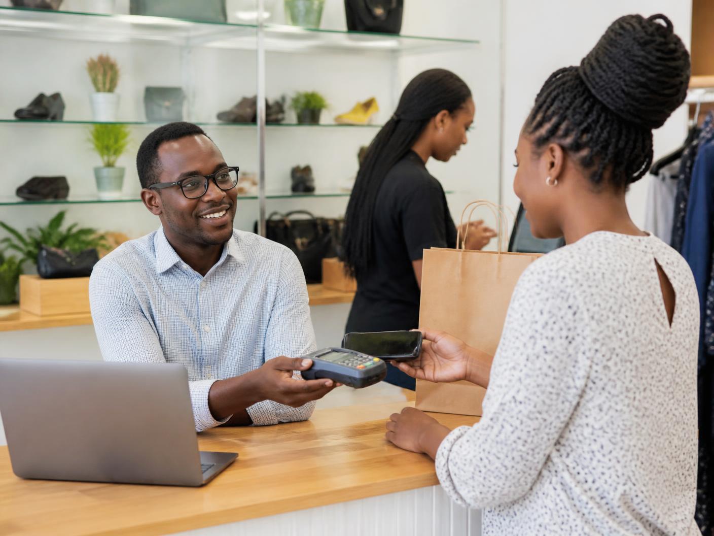 A shop owner in Kenya serving a customer at the counter with a laptop and a mobile payment terminal